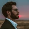 Man with beard and glasses in suit, looking right at a colorful sunset.