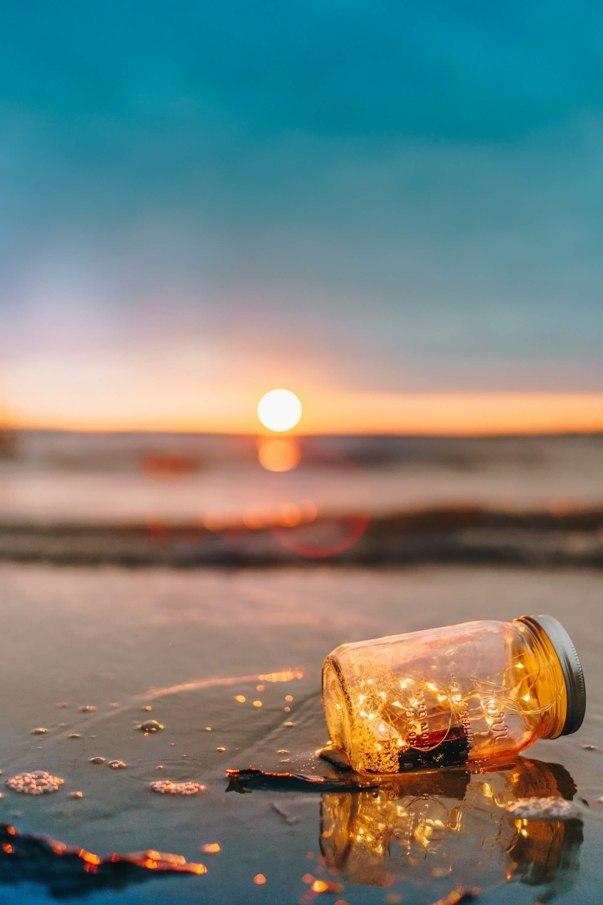 A glass bottle filled with warm string lights rests on a wet beach during sunset.