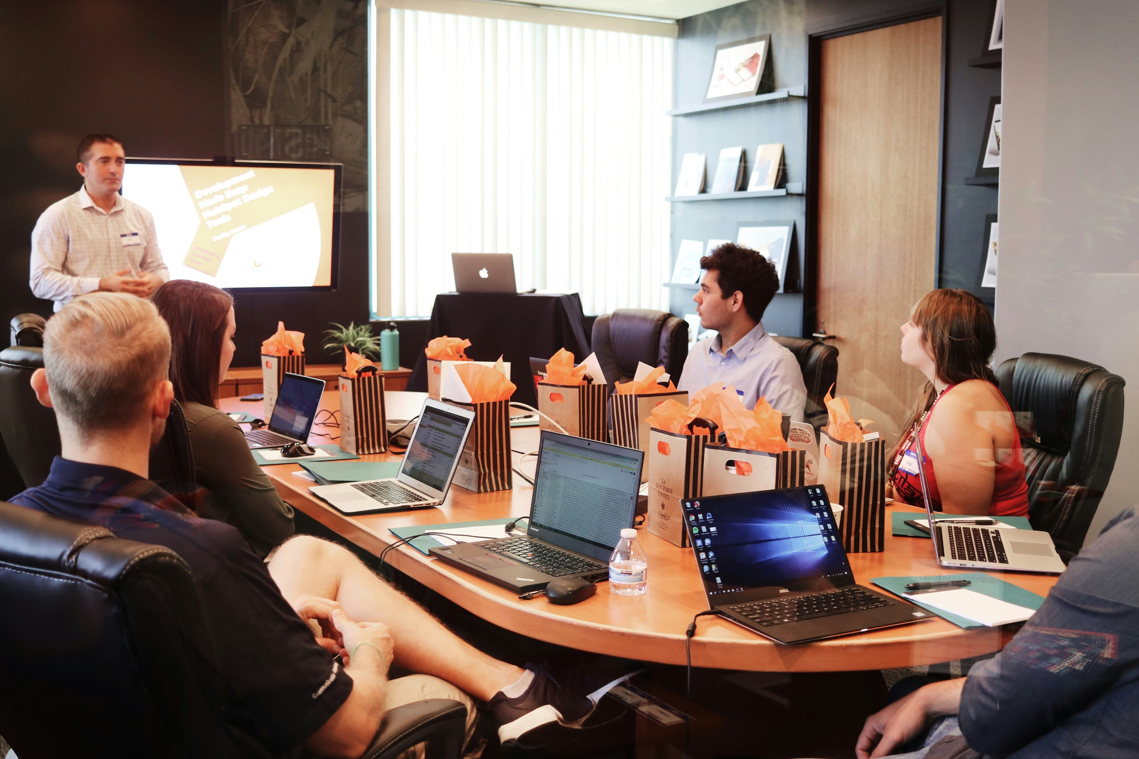 A man presents to colleagues at a conference table with laptops and orange decor.