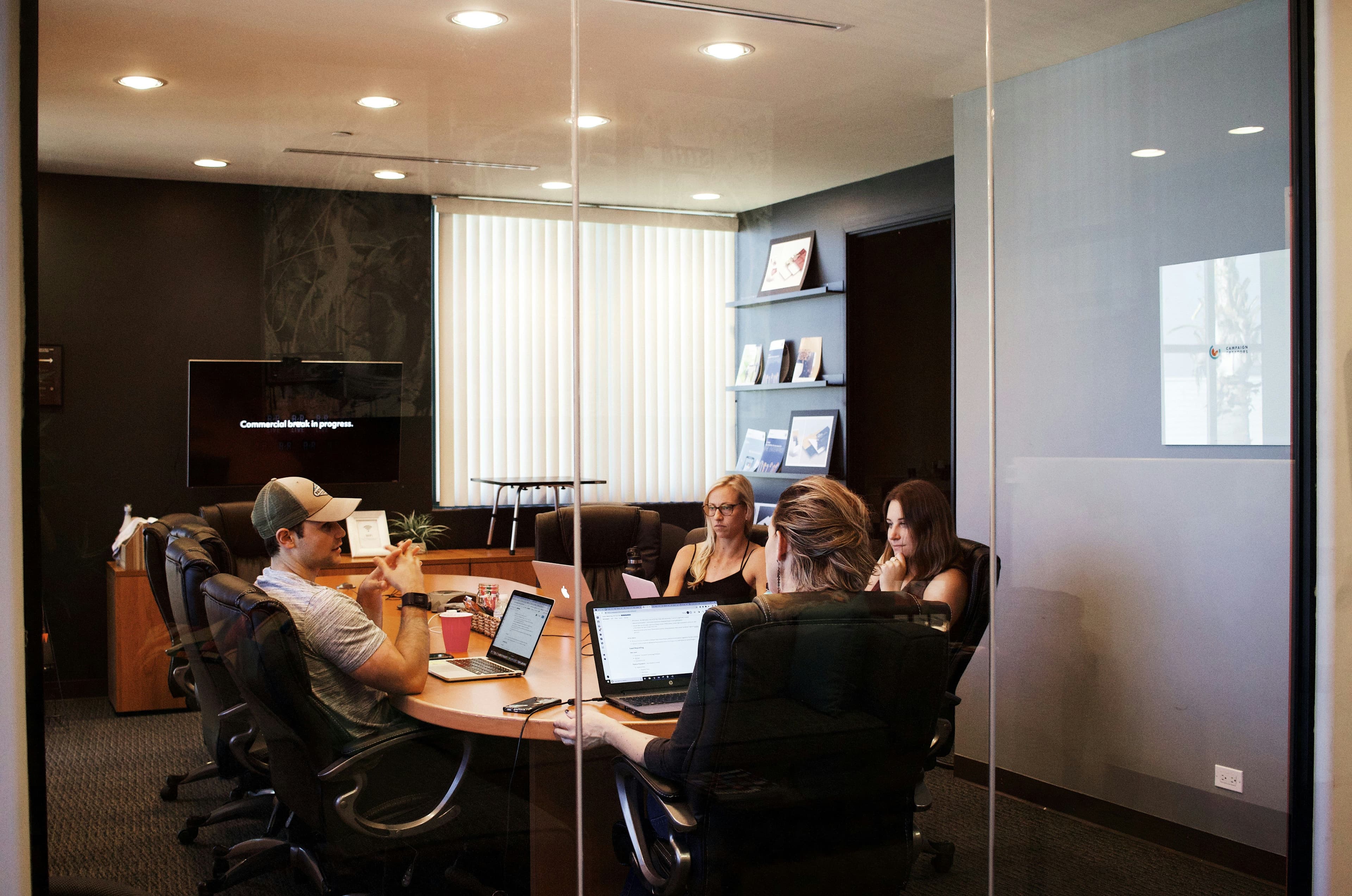 Four people work on laptops around an oval table in a modern glass-walled conference room.