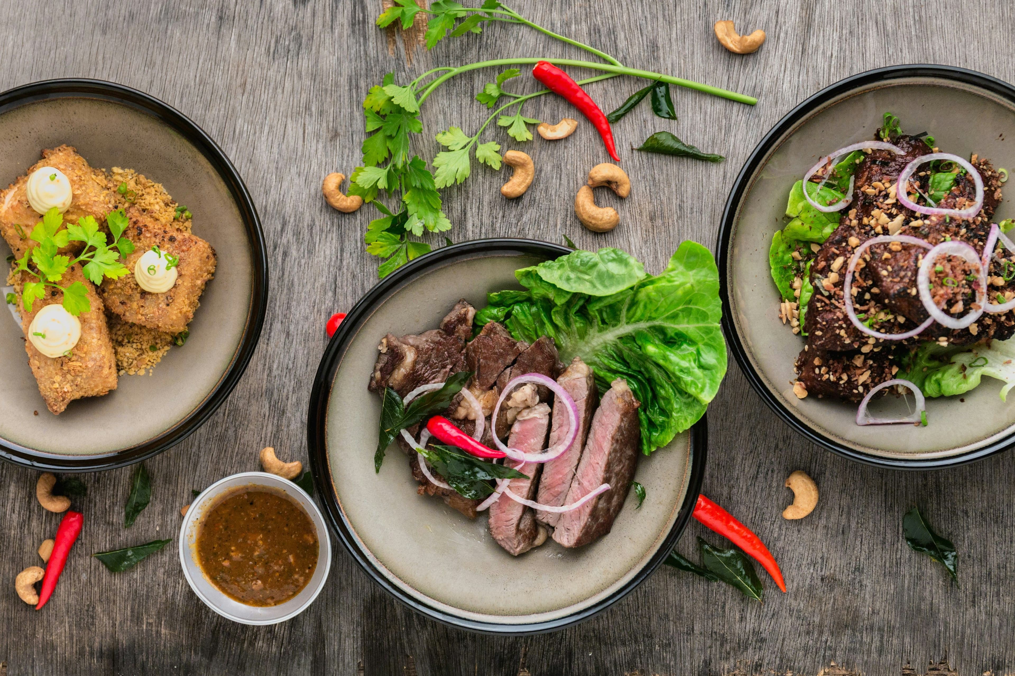 Three gourmet dishes, including meat, tofu, and salad, artfully arranged on a wooden table.